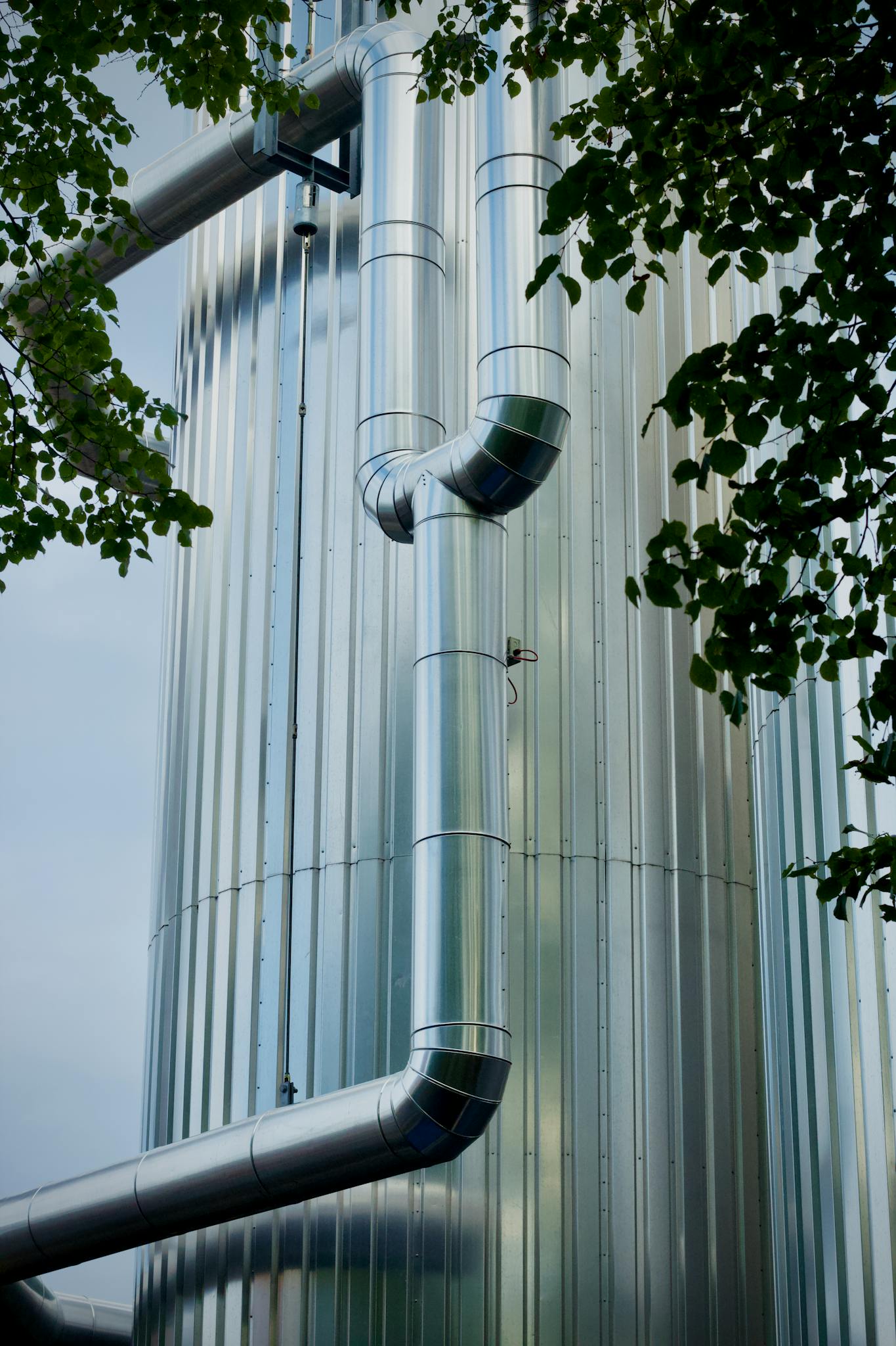View of shiny metal pipes on an industrial tower framed by leaves.